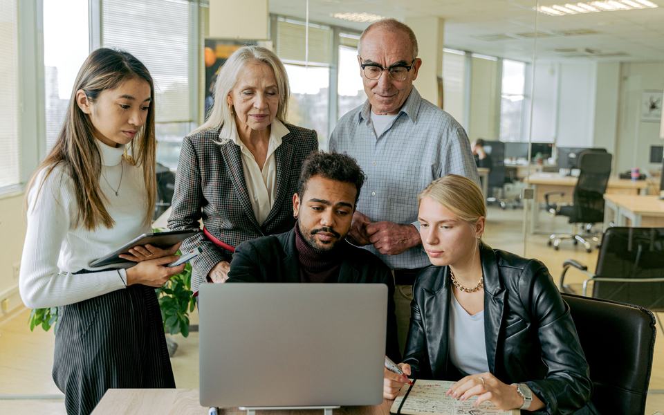 Five colleagues of different ages and backgrounds gathered around a laptop, working through something together