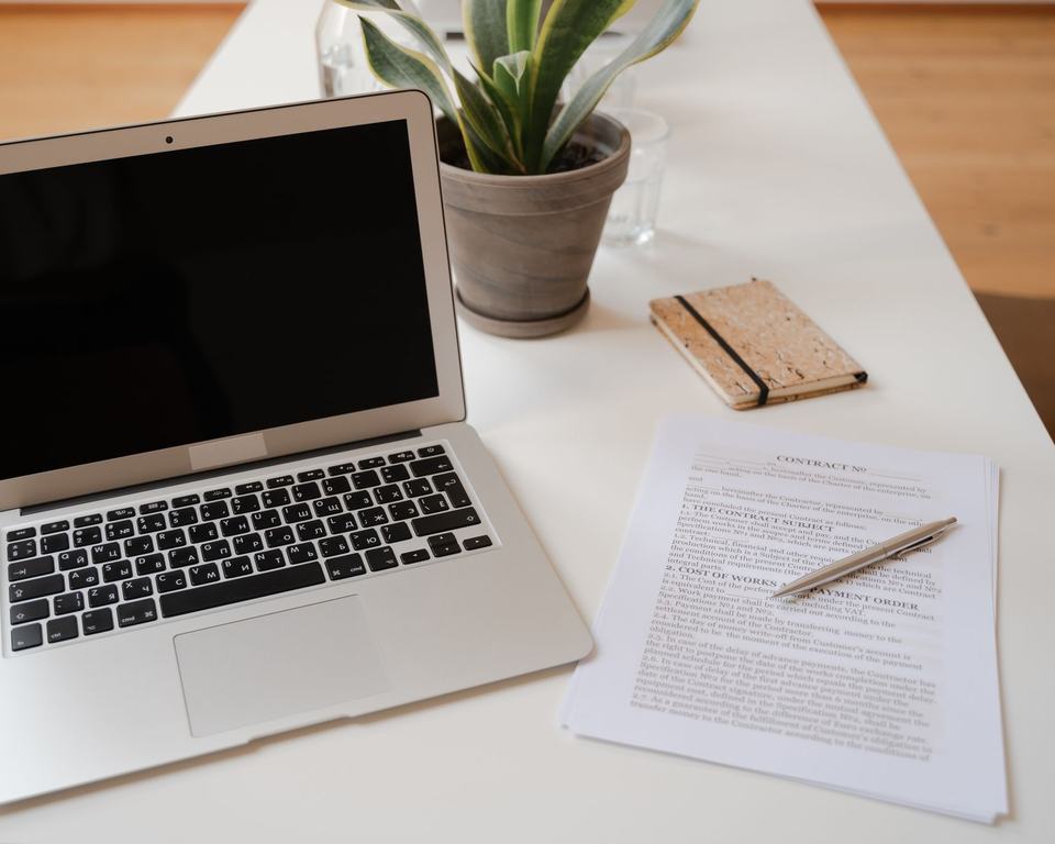 A laptop on a desk beside printed documents and a plant