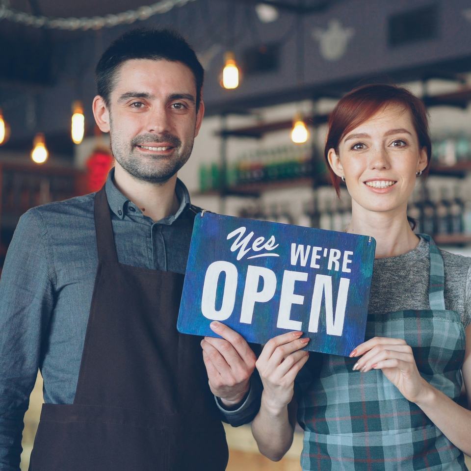 Two smiling café owners in aprons holding a sign that says Yes, we’re open